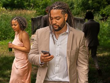 Man in beige suit looking at phone outdoors with woman in pink dress in background.
