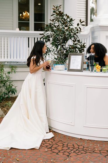 Bride in a white gown chatting at a bar with a bartender outdoors.