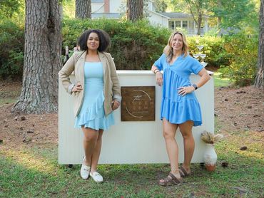 Two women in blue dresses pose by a bar in a wooded outdoor setting.