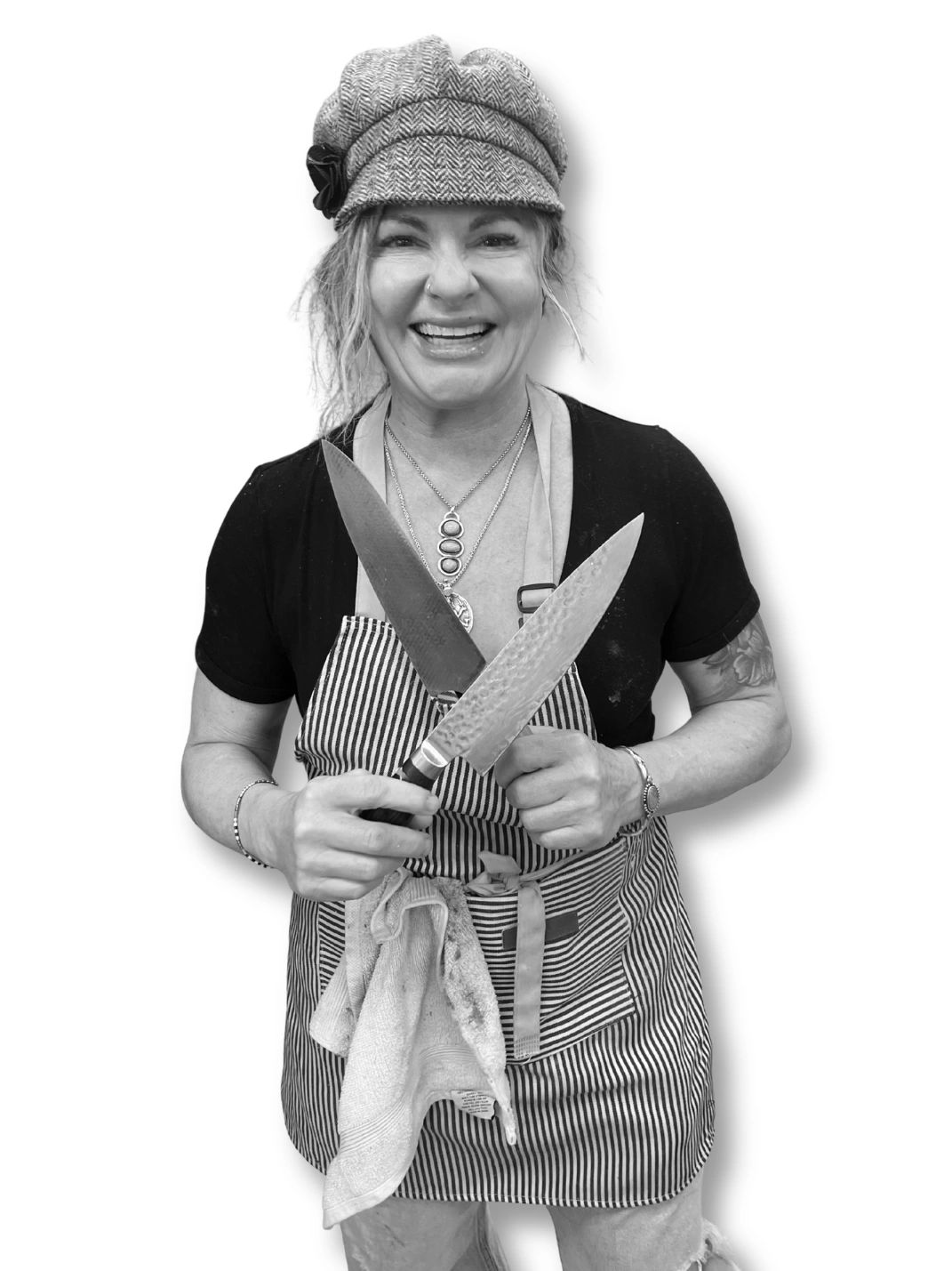 Smiling woman chef holding two large kitchen knives crossed in front of her.