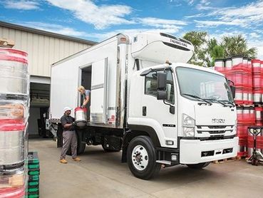 Workers unloading kegs from a white refrigerated truck outside a warehouse.