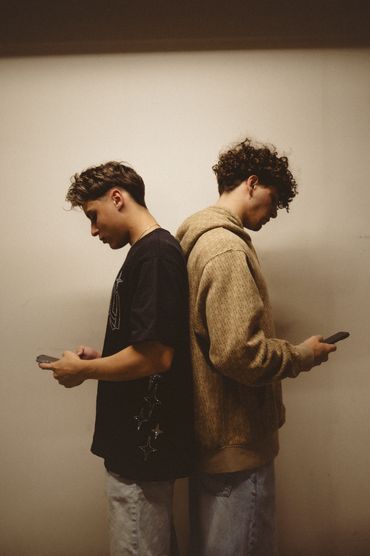 Two young men standing back to back using phones.