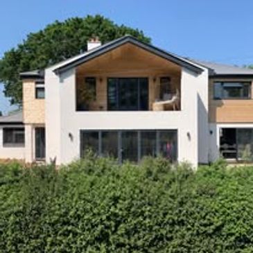 Modern two-story house with large windows and a balcony, surrounded by greenery.