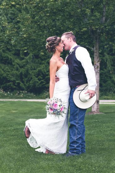 Bride and groom share a moment outdoors. Bride is in a lace dress and groom is holding a cowboy hat.