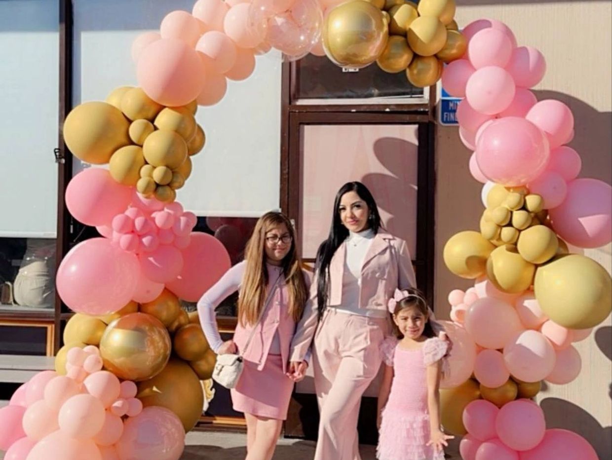 Three females posing under a pink and gold balloon arch outdoors.