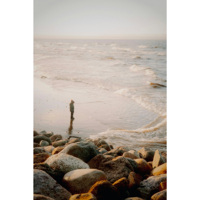 A person stands alone on a rocky beach at sunset, gazing at the waves.
