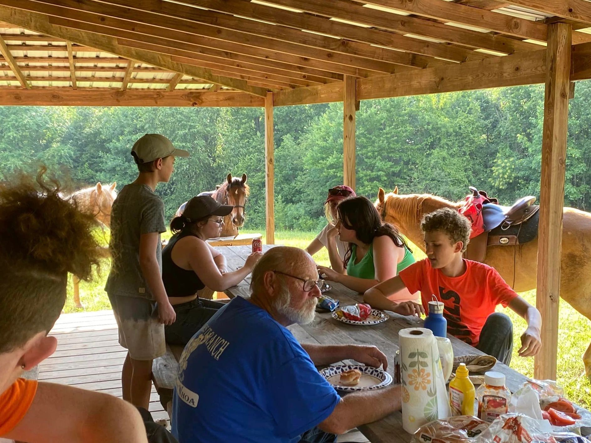 Group eating under wooden shelter with horses nearby.