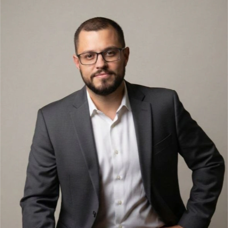 Man with glasses wearing a dark suit and white shirt, posing confidently.