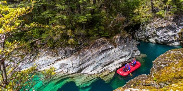 Two people rafting on a clear turquoise river surrounded by rocky cliffs and dense forest.
