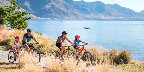 Family cycling by a lake with mountains in the background.