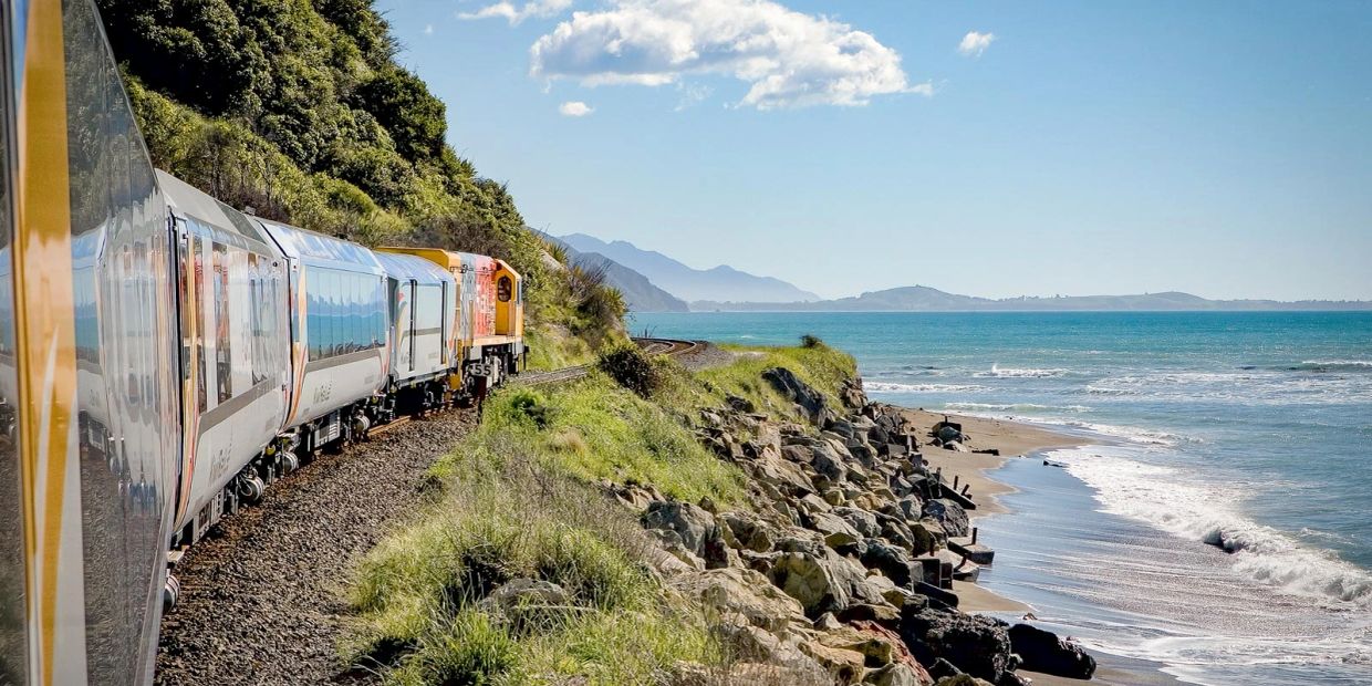 Train traveling along a coastal track with ocean waves and mountains in the background.