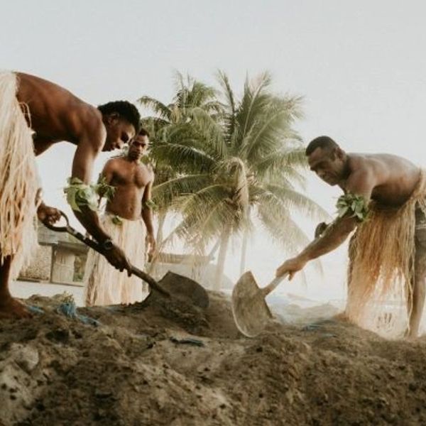 Three men in traditional attire digging with shovels near palm trees.