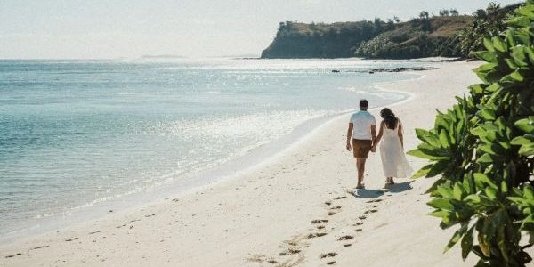 A couple walks hand in hand on a sunny beach with footprints in the sand.