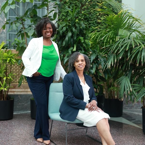 Two professional women posing in a lush indoor garden setting.