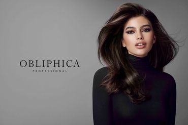 A stylish woman with flowing hair and smoky eye makeup poses in a black turtleneck against a gray backdrop.