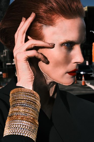 Elegant person with red hair and blue eyes adorned with multiple sparkling gold bracelets.