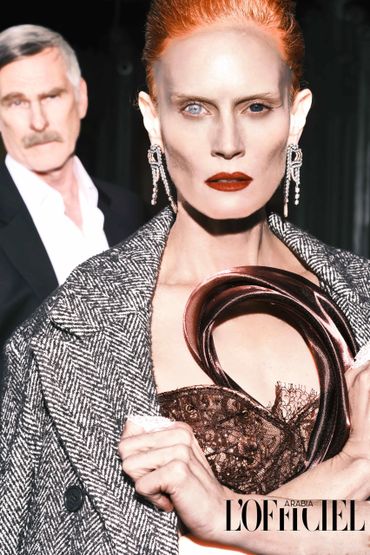 Striking fashion portrait with bold makeup and textured coat, featuring a man blurred in the background.