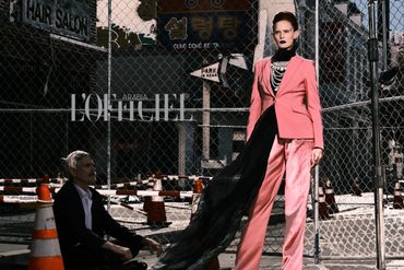 Model in a striking pink suit with black veil poses against a chain-link fence in an urban setting.