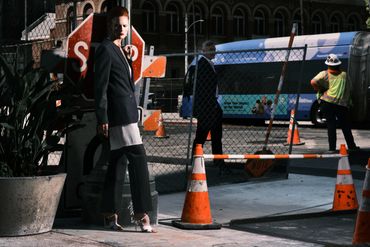 A stylish woman stands near a construction site with orange cones and a stop sign.