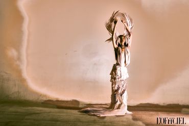Model in an ethereal dress with feathered headpiece poses against a soft, muted backdrop.