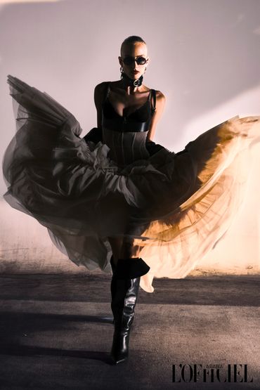 Fashion model in dramatic black outfit and flowing skirt against a plain backdrop.