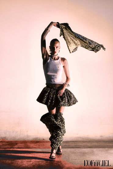 Model in a chic white tank top and textured skirt with matching leg warmers, striking a dynamic pose with a scarf.
