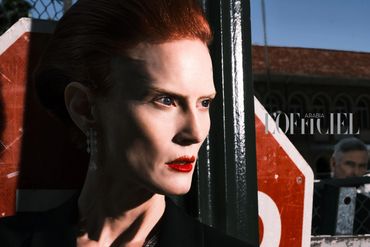 Close-up of a red-haired woman with red lipstick, posing near a stop sign and a fence.