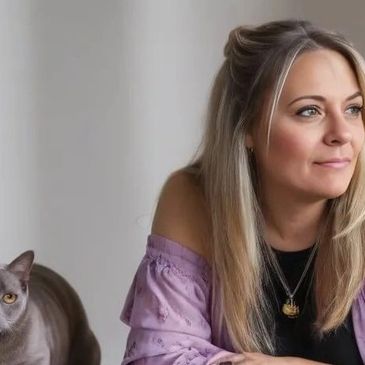 A woman with blonde hair looks thoughtfully to the side with a gray cat beside her.