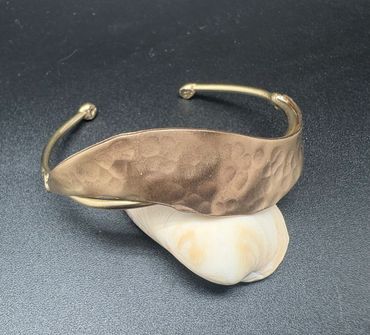 Hammered brass cuff bracelet resting on a seashell against a dark background.