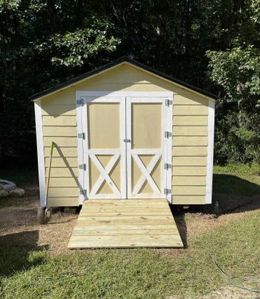 A yellow and white garden shed with a wooden ramp and a shovel leaning against it.