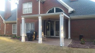 Covered patio with grill and chair on a brick house's backyard.