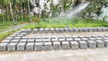 Concrete bricks curing under water sprinkler in a green outdoor setting.