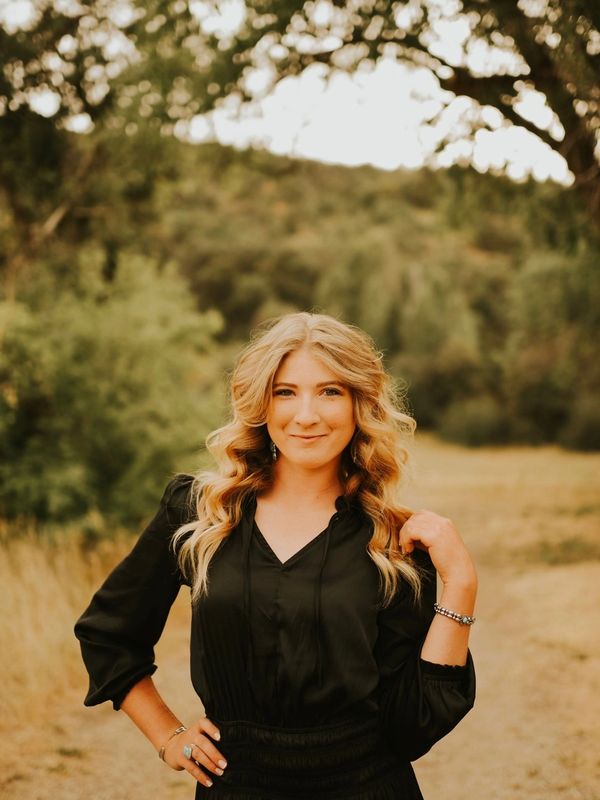 Young woman with wavy blonde hair in a black dress smiles outdoors.