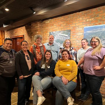 Group of nine people posing indoors against a brick wall with framed aerial photos.
