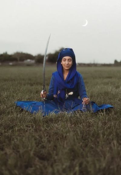 Woman in blue meditating with a sword in a grassy field under a crescent moon.