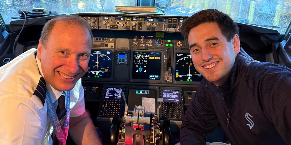 Two men smiling inside an airplane cockpit with controls and displays.