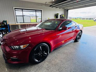 Shiny red sports car parked in a spacious garage with open door showing a green lawn outside.