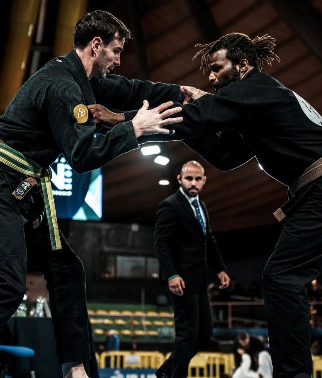 Two martial artists grappling in a Brazilian Jiu-Jitsu match with a referee observing.