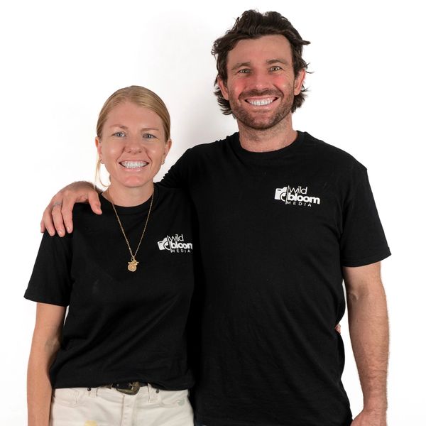 Two smiling people wearing black Wild Bloom Media t-shirts pose together against a white background.