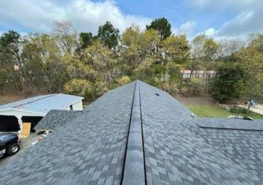 View of a gray shingled rooftop with trees and a building in the background.