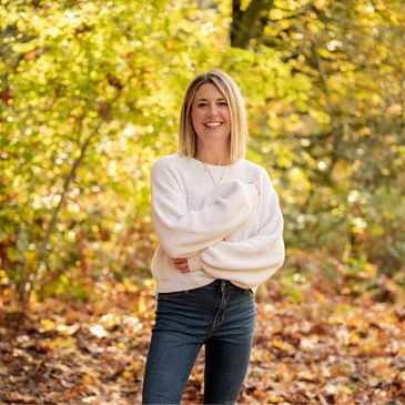 Smiling woman in white sweater stands in autumn forest with crossed arms.