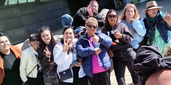 A group of diverse women posing outdoors, some making peace signs and thumbs up gestures.