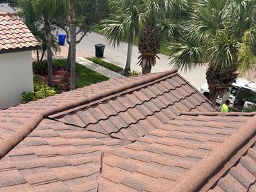 Brown tiled roof with visible damage under sunlight and palm trees in the background.
