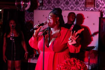 A woman in a red dress passionately sings on stage with band members behind her.