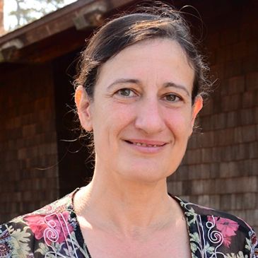A woman smiling outdoors in front of a wooden building.