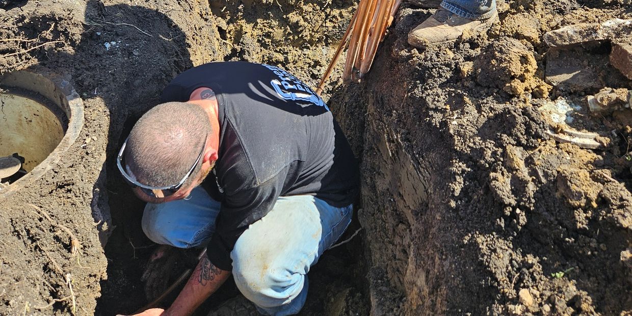 A man working in a deep trench, handling copper pipes in muddy soil.