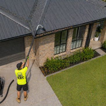 Worker cleaning gutters on a house using a long pole.