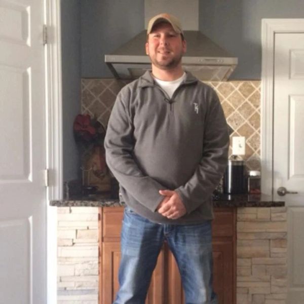 Man smiling in a kitchen, wearing a cap and casual clothes.