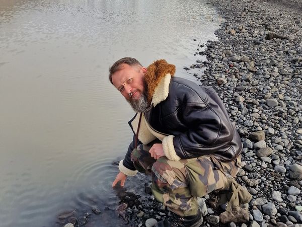 Man in leather jacket crouching by a rocky riverbank, touching the water.