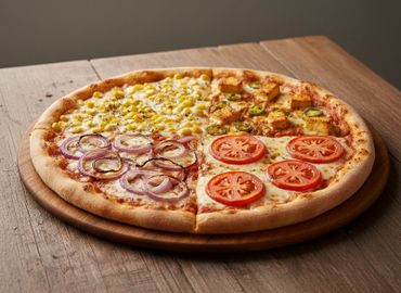 A four-flavor pizza with corn, paneer, tomato, and onion toppings on a wooden table.